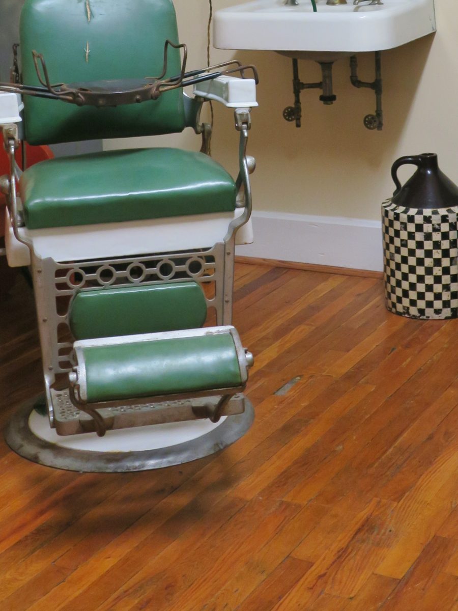 A green barber chair beside a white porcelain sink on a wooden floor.