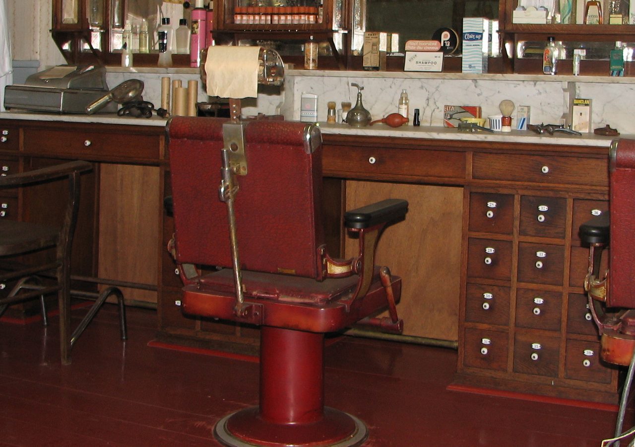 A barbershop interior with a red upholstered chair facing a wooden cabinet and a wall mirror.
