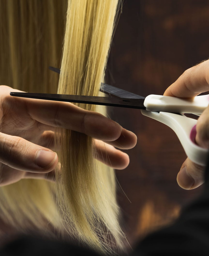 Two hands lift a section of long hair over a comb and cut it with shears. Only the hands, the hair and the tools are in frame.