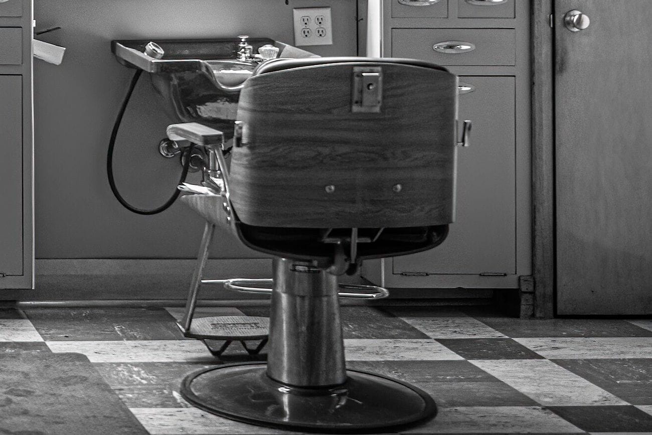 A black and white view of an empty barber chair in front of a wall basin and wooden cabinets, on a chequered floor.