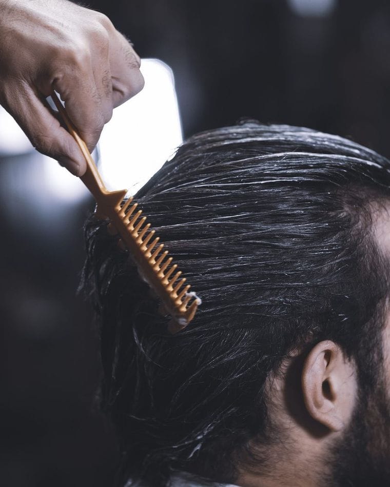 A barber draws a wooden comb back through a client's slicked dark hair, seen from the side with the face turned away.