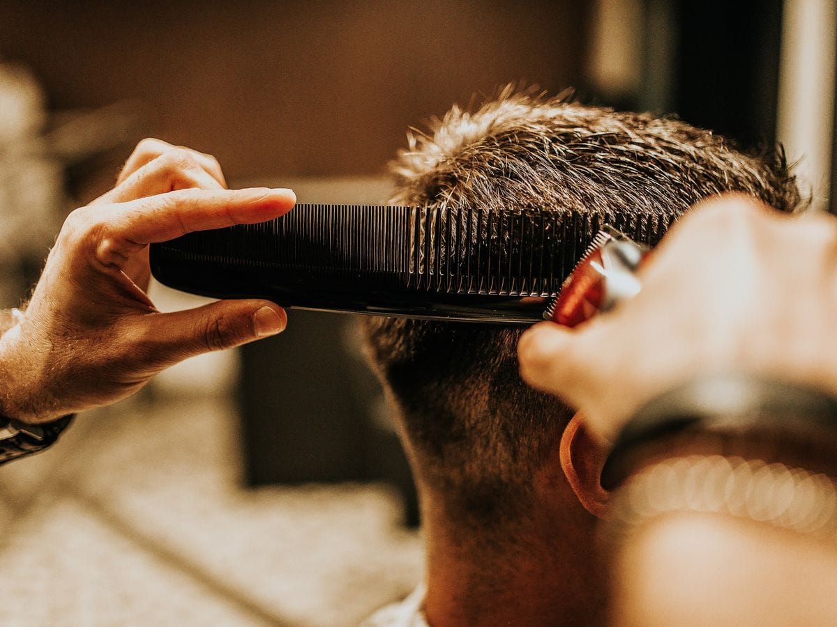 A barber holds a black comb against the back of a client's head and follows it with a red clipper, the client seen from behind in a warmly lit shop.