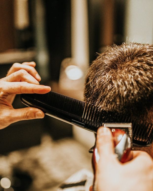 A barber holds a black comb against the side of a client's head while a clipper follows beneath it, the client seen from behind.