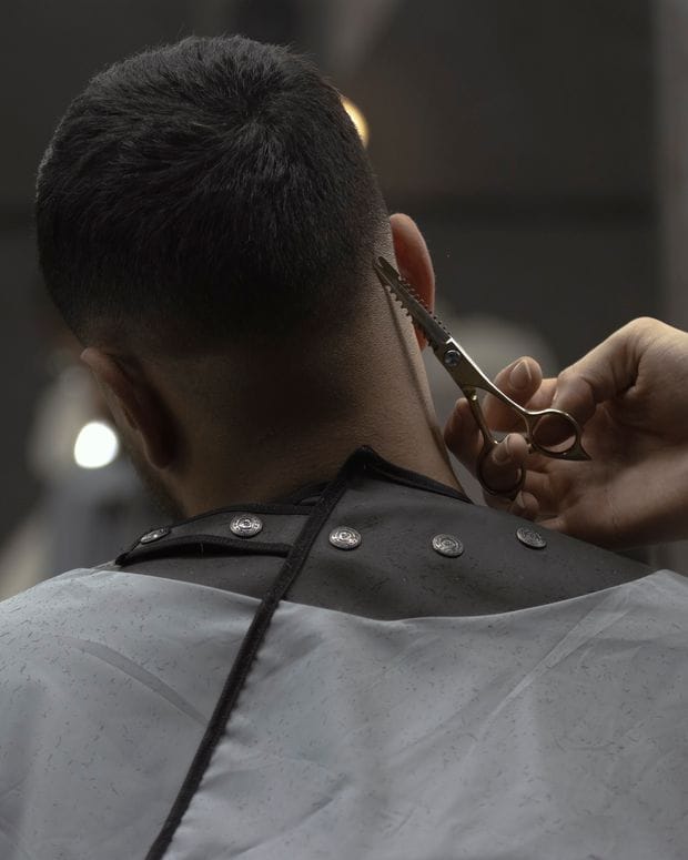 A barber trims the hair at the nape of a caped client with small scissors, the client seen from behind.