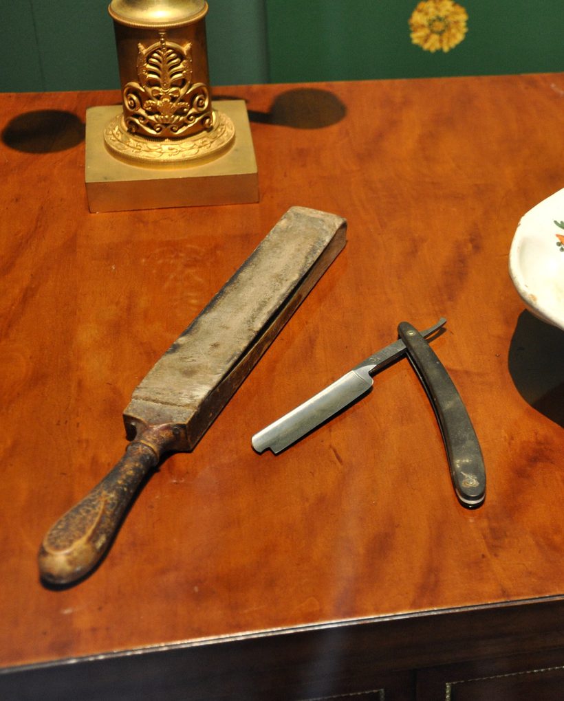 A strop paddle and open straight razor on a polished table beside a shaving basin