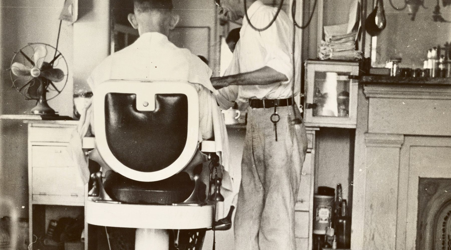 An archive photograph of a caped client in a cast-iron chair, the barber mixing lather at the counter behind