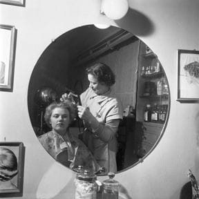 A hairdresser setting a client's hair, reflected in a round salon mirror.