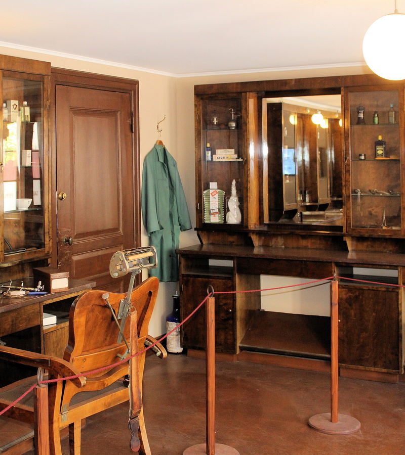 A museum hairdressing room with dark wooden cabinetry and a mirrored cabinet behind a rope barrier.