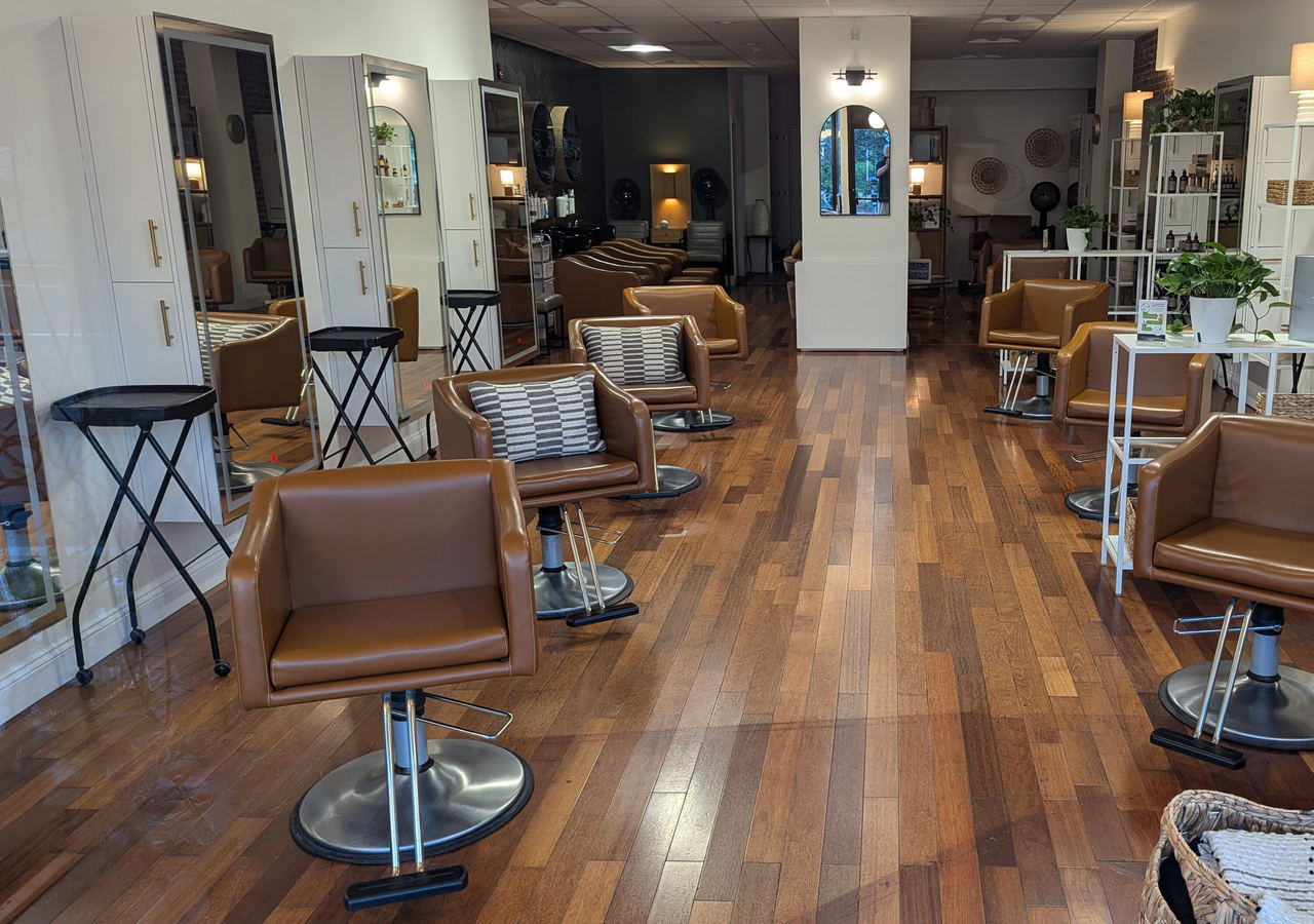 A hair salon with tan leather chairs on a wood floor, facing tall mirrors and open shelving.