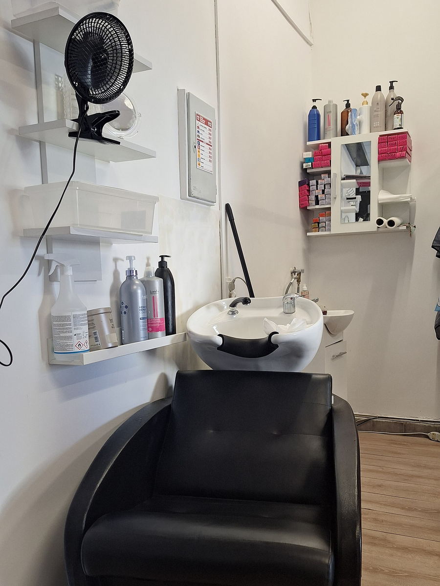 A washing station with a white basin, a black reclining chair and wall shelves of bottles.