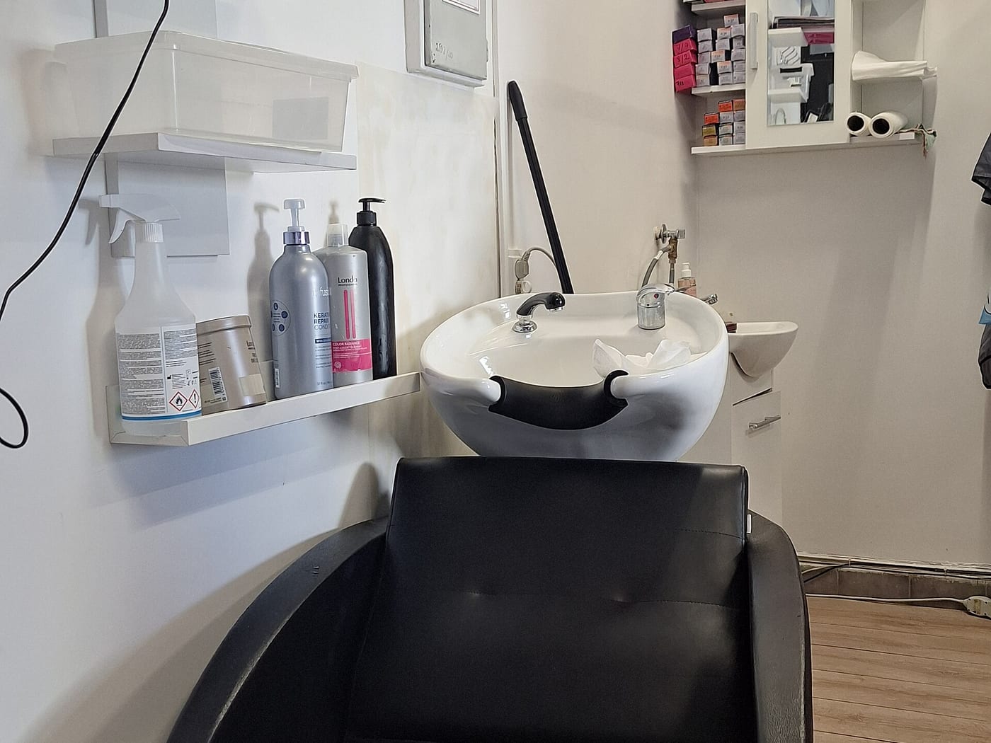 A backwash basin with a black reclining chair and shampoo bottles on a white shelf.