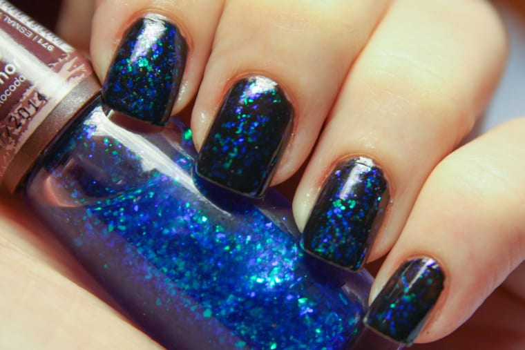 A hand with short nails painted in dense blue glitter polish, resting on a pale surface.