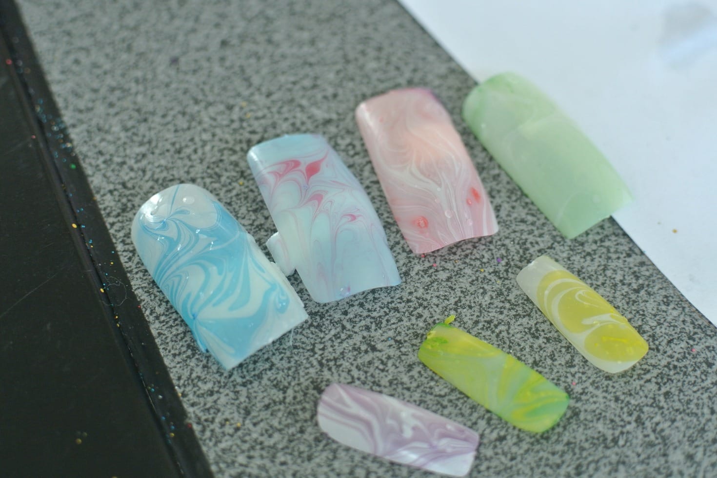 Practice nail tips painted in pastel marbled colours, laid out on a grey table.