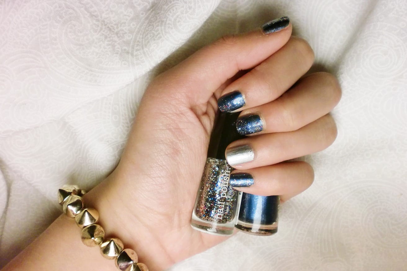 A hand with metallic silver and dark blue nails holding a small bottle of silver polish.