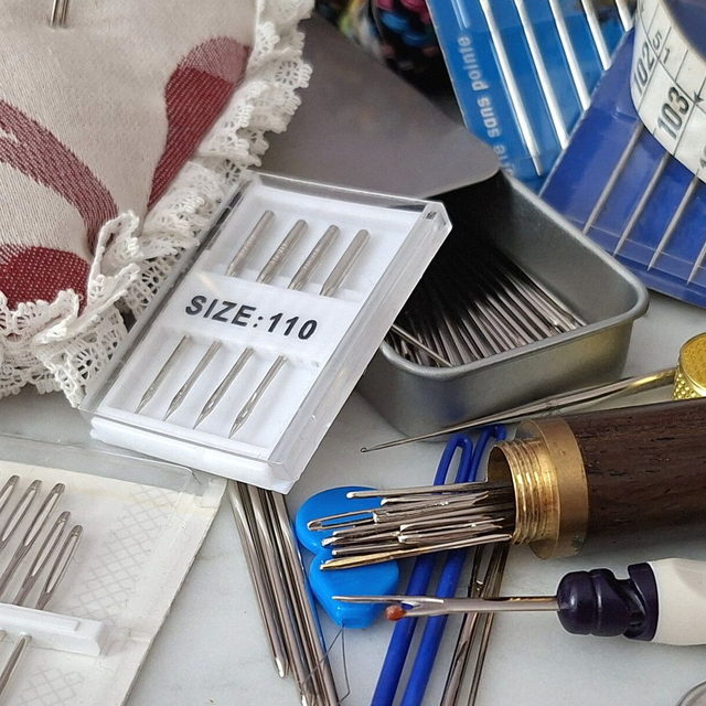 A closer crop of the same workbench: the eyes of three hand sewing needles beside the end of the tape measure.