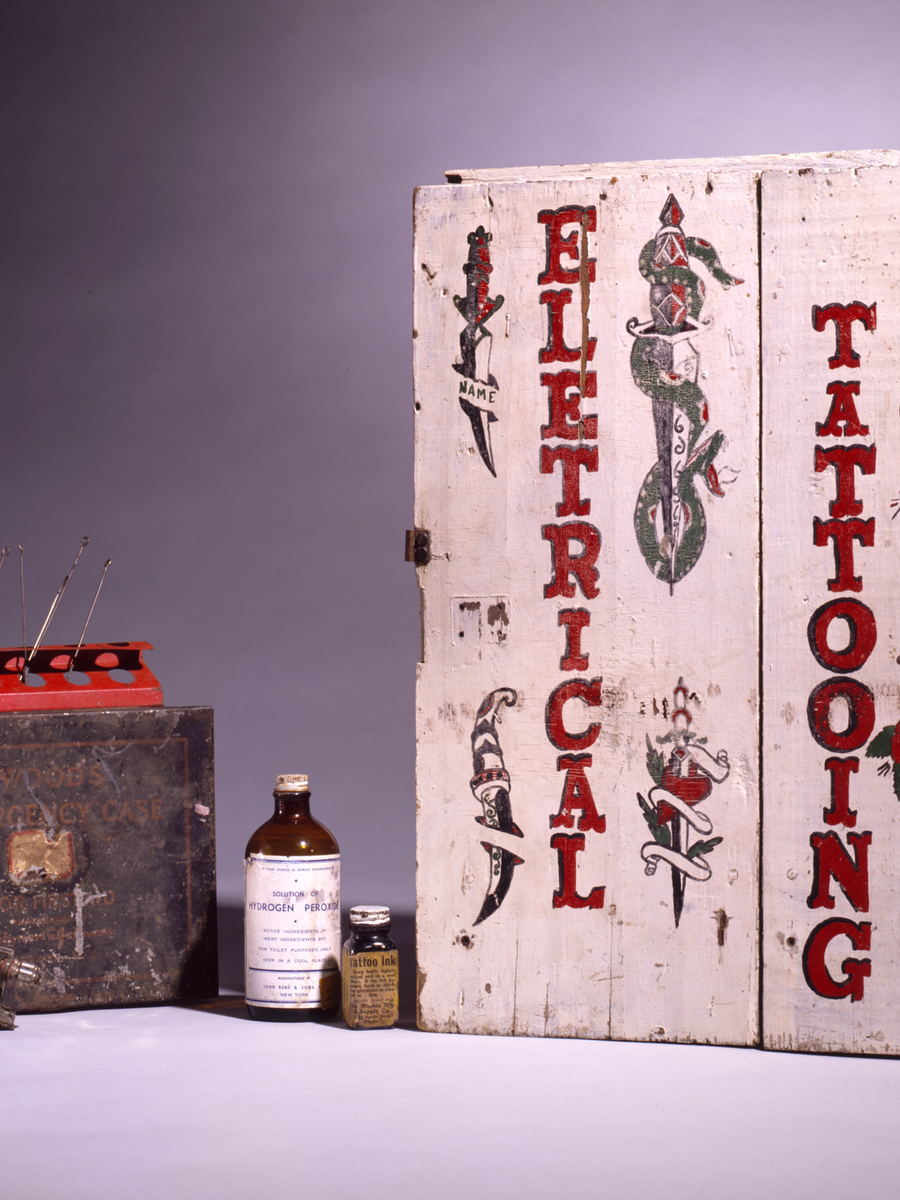A painted tattooist's case standing open: a machine, needle bars and ink bottles held in fitted compartments.