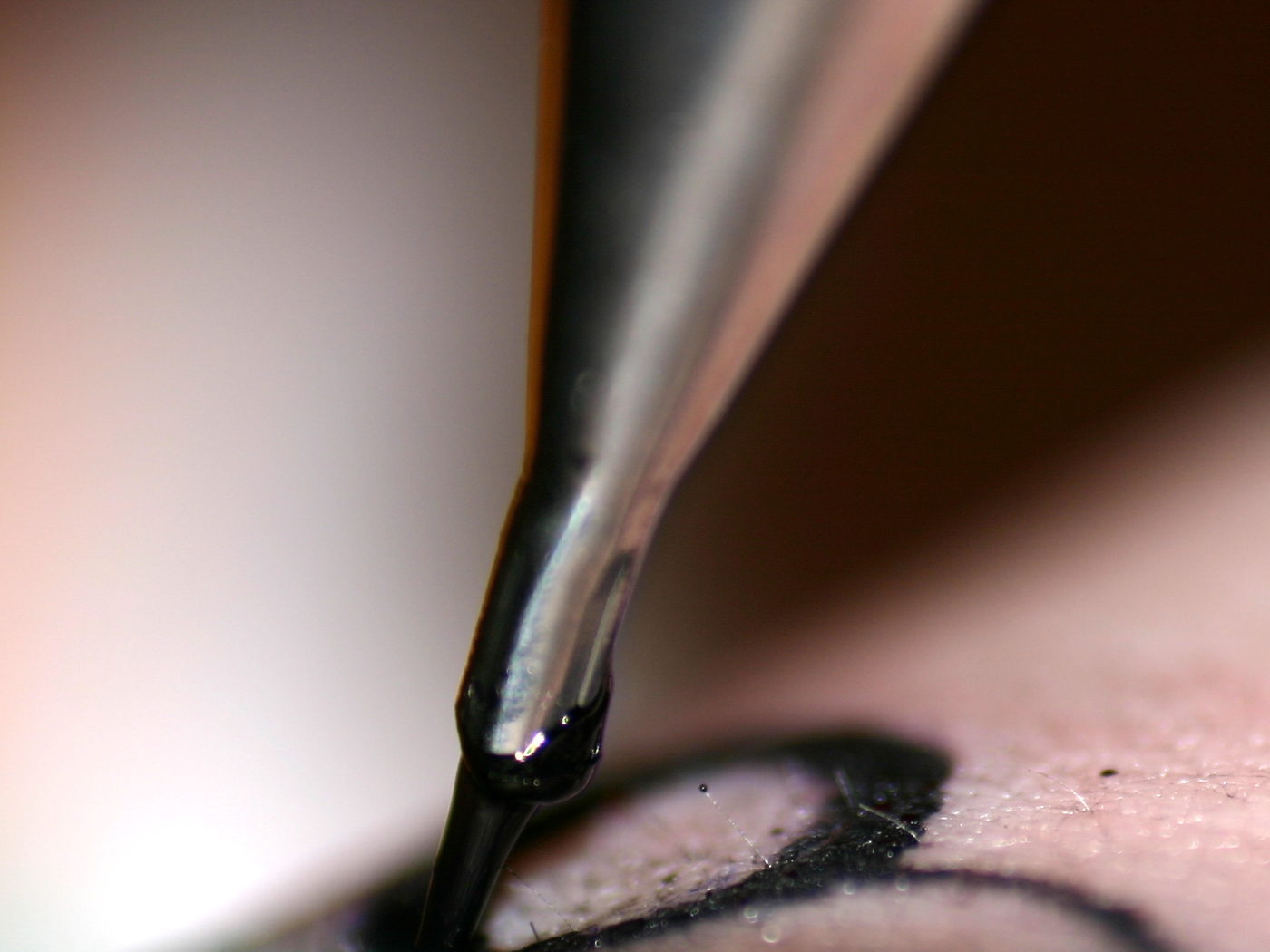 A machine needle held close to the skin, ink gathering at the tip beside a line already drawn.