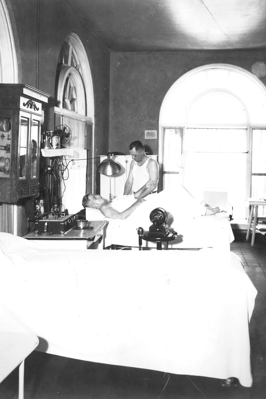 A black-and-white archive photograph of a bathhouse massage room, with a masseur working beside a client on a padded table under a tall arched window.