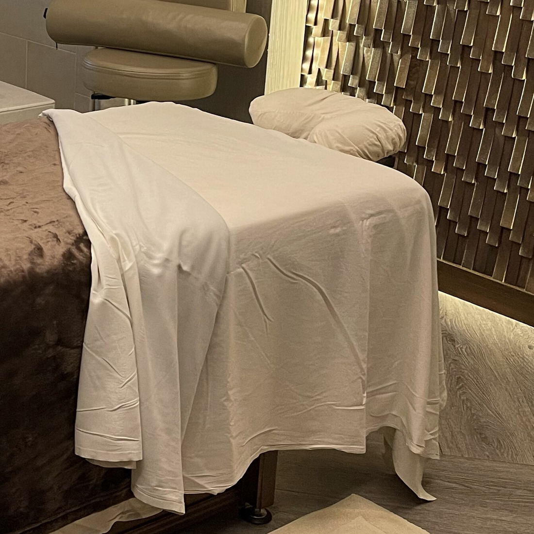 A white sheet over a massage table with a face cradle and a cushioned arm, beside a textured wood-tile wall.