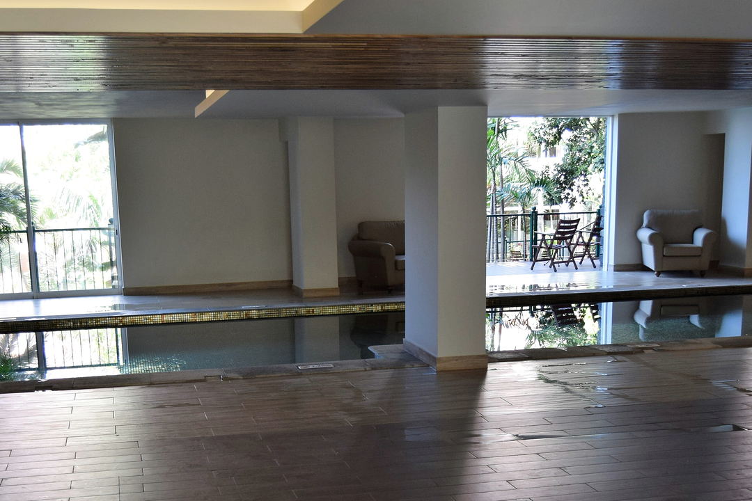 An indoor pool hall with a white column, glass doors onto greenery, a balcony above and armchairs along the side.