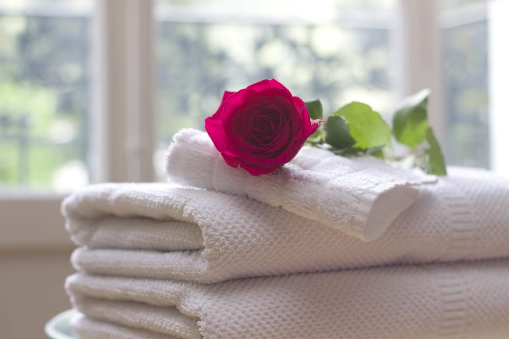 A stack of folded white towels on a sunlit ledge with a single red rose laid on top.