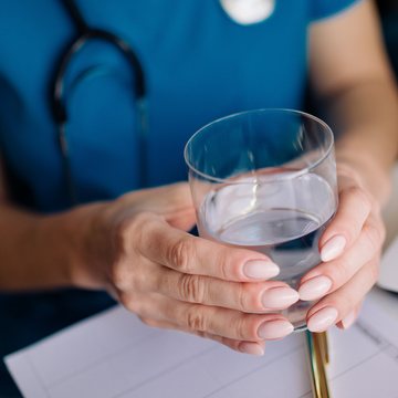 A clinician in scrubs holding a glass of water above a notepad
