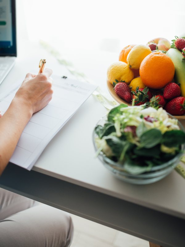 A hand writing in a notebook beside a bowl of oranges, lemons and strawberries