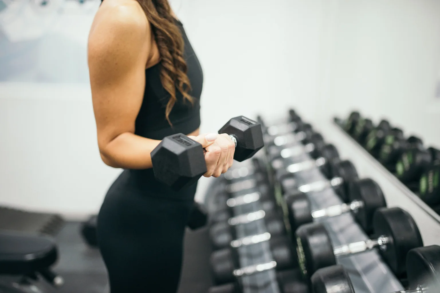 Member curling a dumbbell beside a loaded rack, photographed without a face in frame