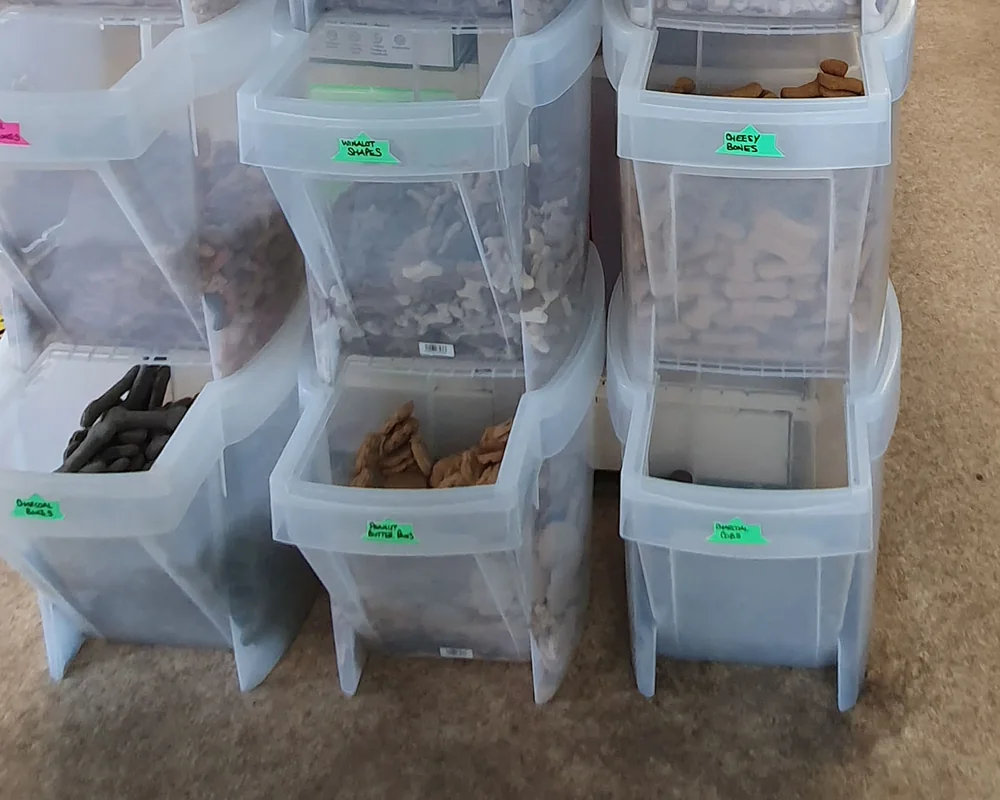 Stacked clear gravity bins of loose dog biscuits with handwritten flavour labels.