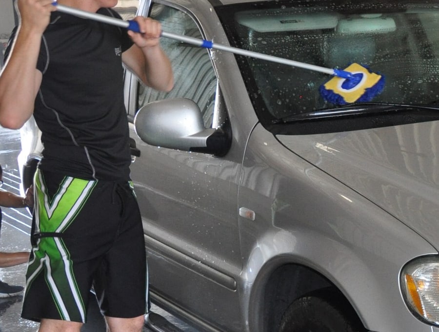 A wash mitt on a long handle drawn across the windscreen of a soapy silver SUV, washed by hand