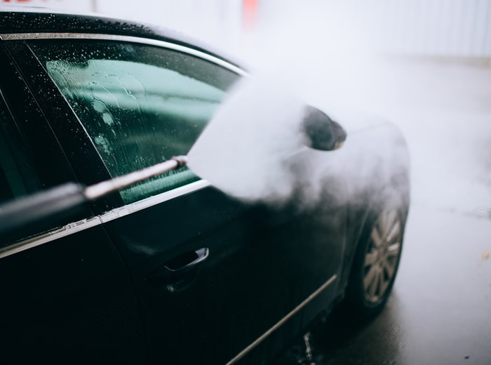 A pressure lance laying foam down the flank of a black car, glass and paint beaded with water