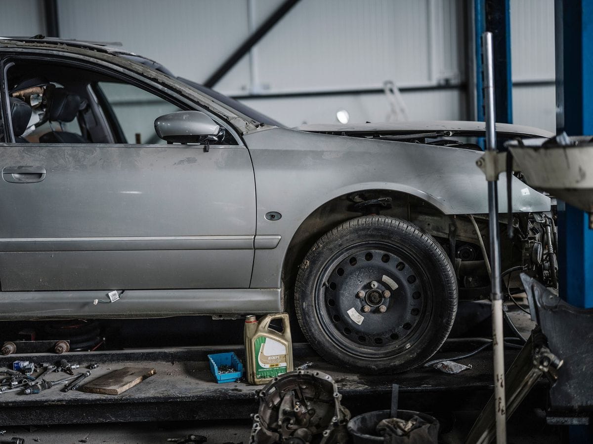 A partially dismantled car raised on a lift in a repair shop, with tools and parts laid out on the floor beneath it.