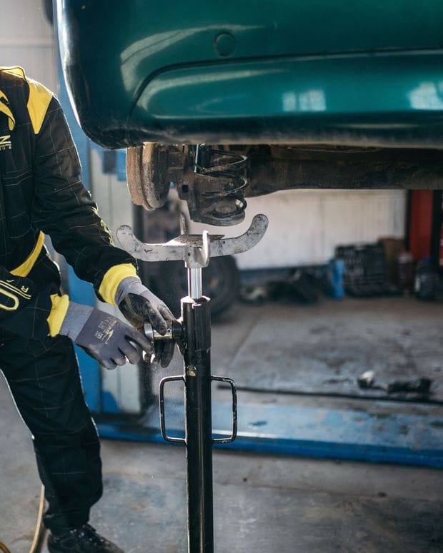 The underside of a car raised on a lift in a repair shop, with a technician working on the front suspension.