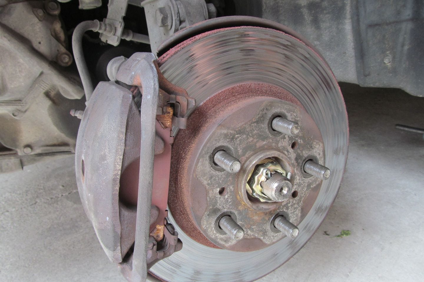 A brake disc and caliper on a wheel hub with the wheel removed, the studs and flexible hose visible.