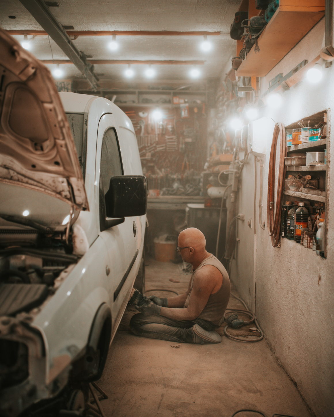A figure at floor level preparing a vehicle panel in a wide workshop.