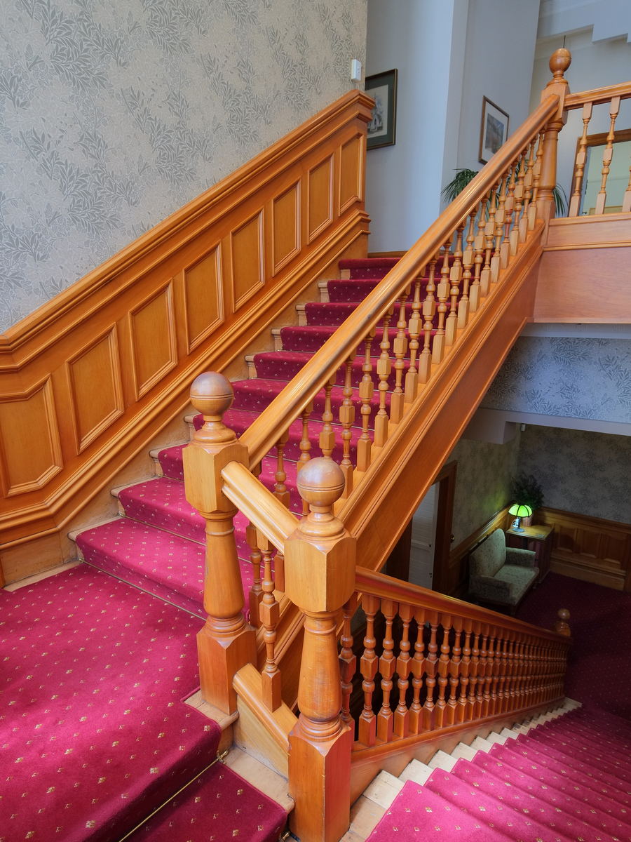 A broad timber staircase turning at a half landing, with a carved newel post and a tall window above it.