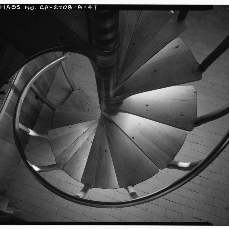 A spiral staircase photographed from above, the treads winding down into shadow around a plain iron newel.