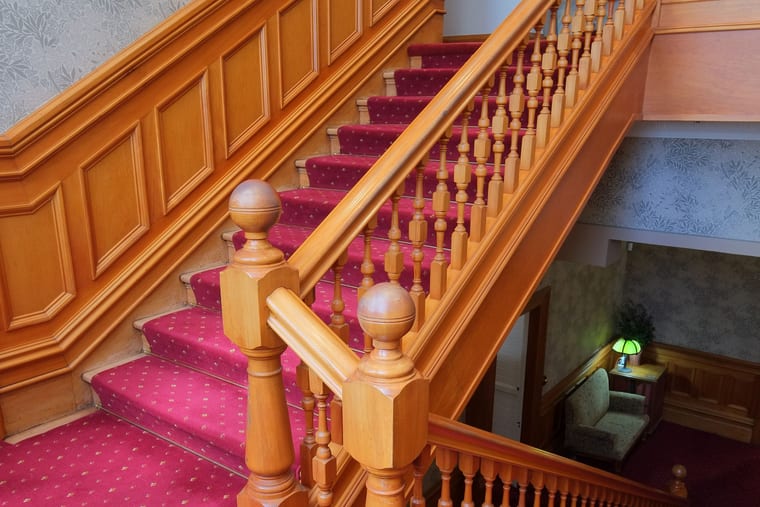 A varnished timber staircase with turned balusters and a patterned red carpet over the treads.