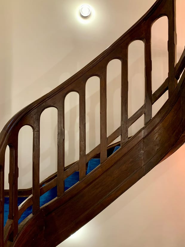 A curved timber balustrade rising past a plain wall, with a ceiling light above it.