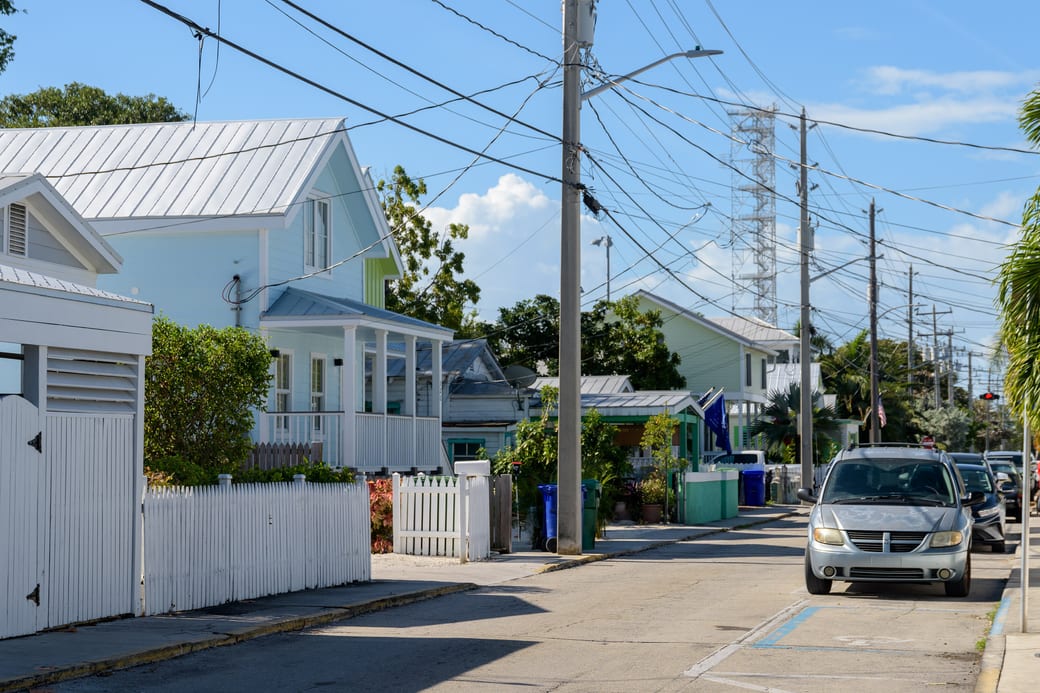 A residential street of pale clapboard houses behind white picket fences, with palms along the kerb.