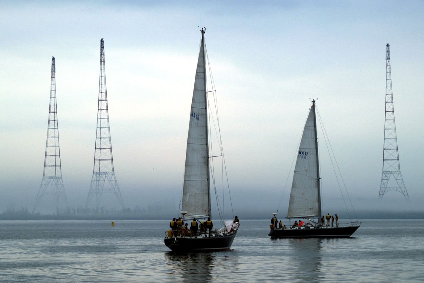 Two sailing yachts with crews aboard on calm grey water, with tall lattice pylons standing in the haze behind.
