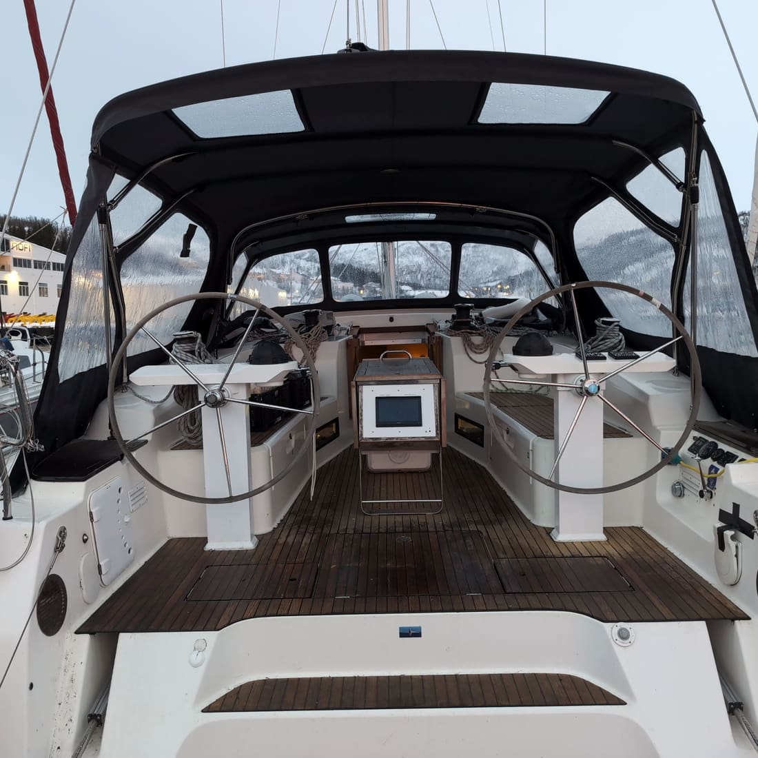 The wheel cockpit of a sailing yacht under a dark bimini, with a teak sole, moored in a marina.