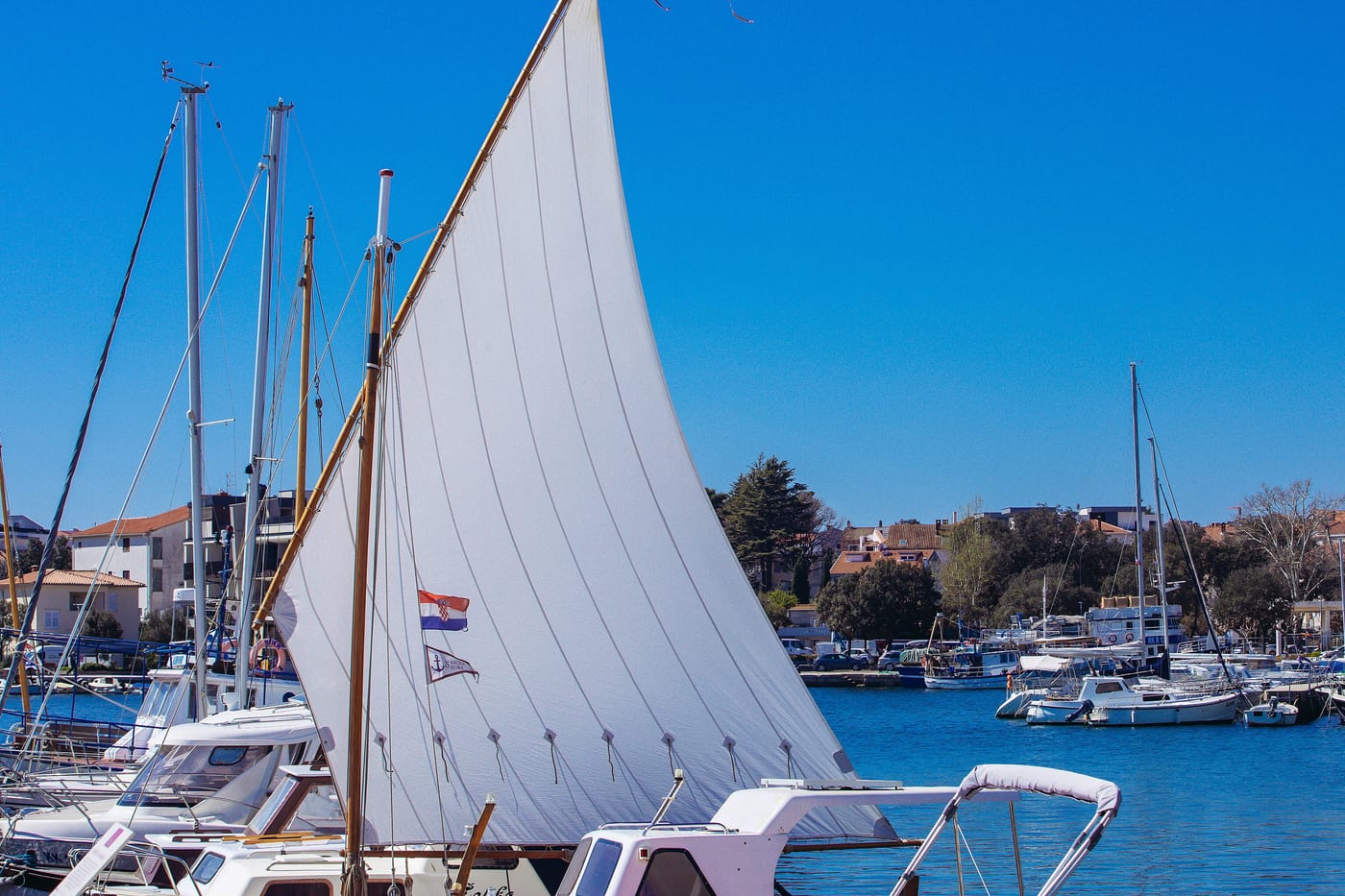 A white lateen sail raised on a small wooden boat inside a harbour of moored yachts, under a clear blue sky.