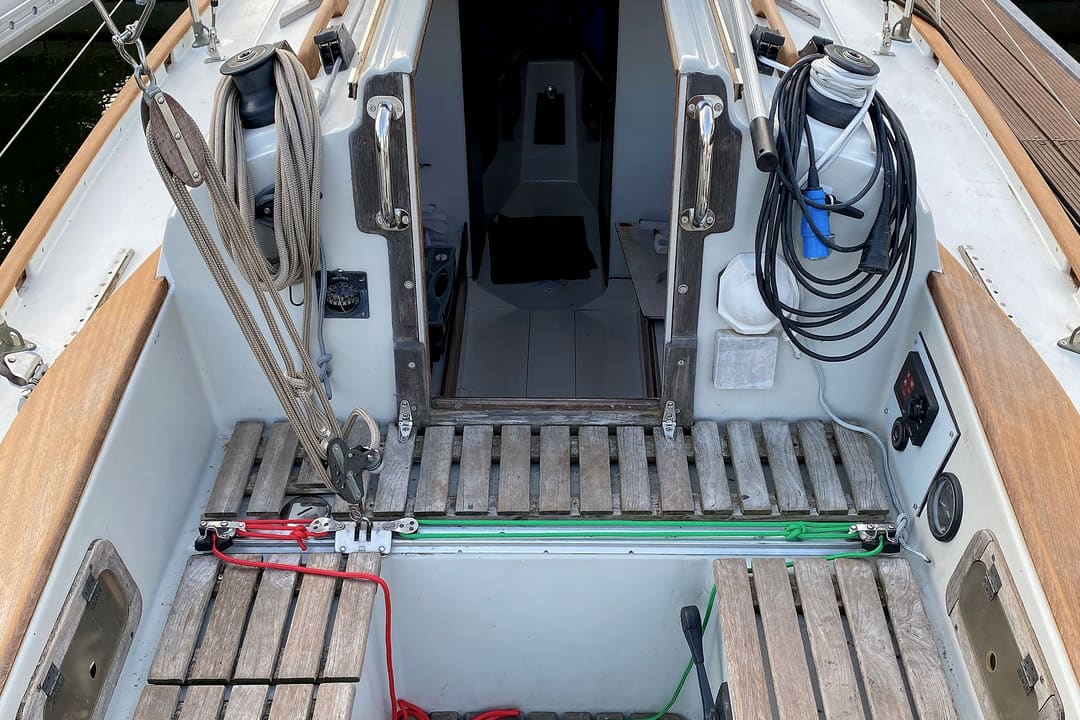 The cockpit of a small sailing yacht seen from the stern, with teak slatted seats, winches and the open companionway.