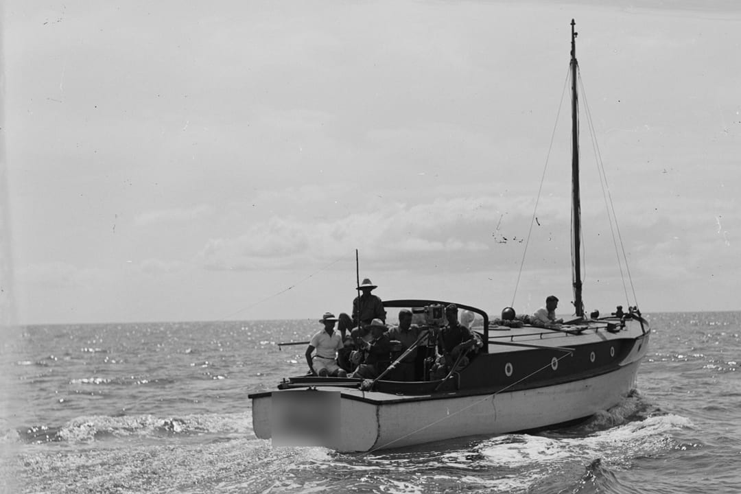 An archive black-and-white photograph of a wooden yacht under way in open water, seen from astern with a small party aboard.