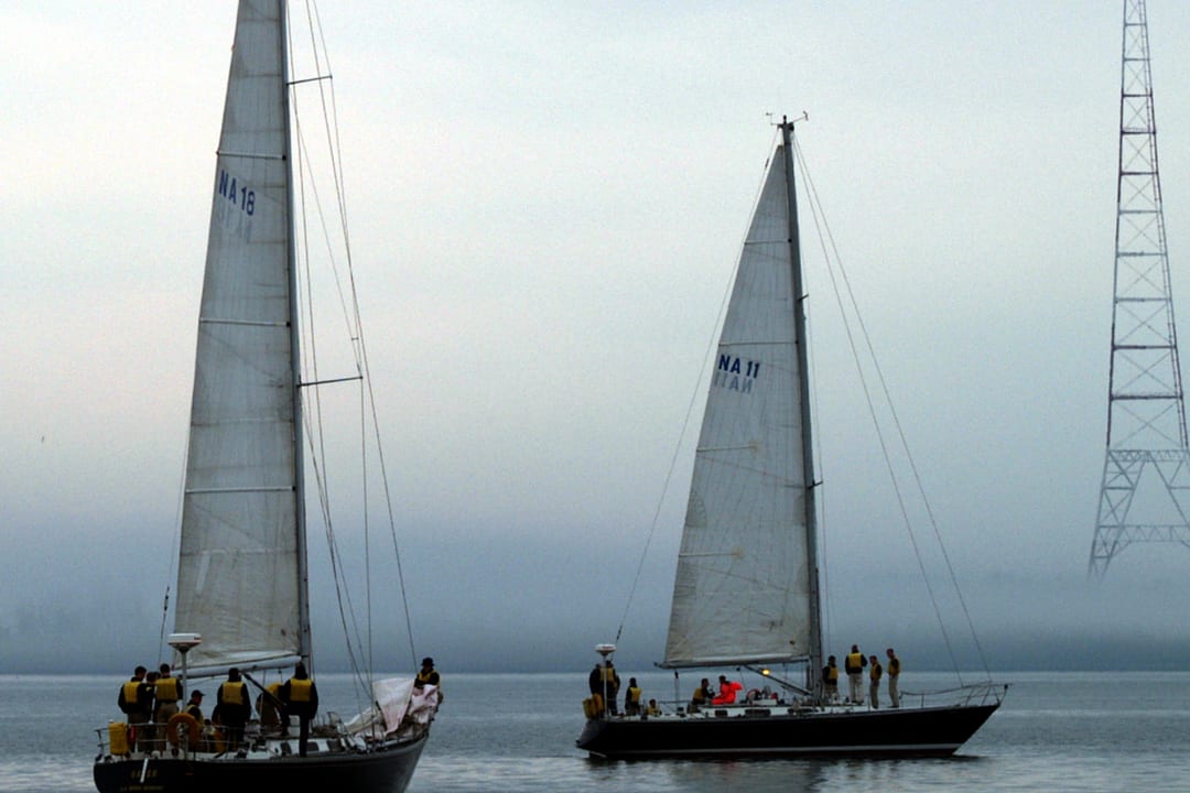 Two white sailing yachts with their mainsails up, lying off a grey misty shore with crews on deck.