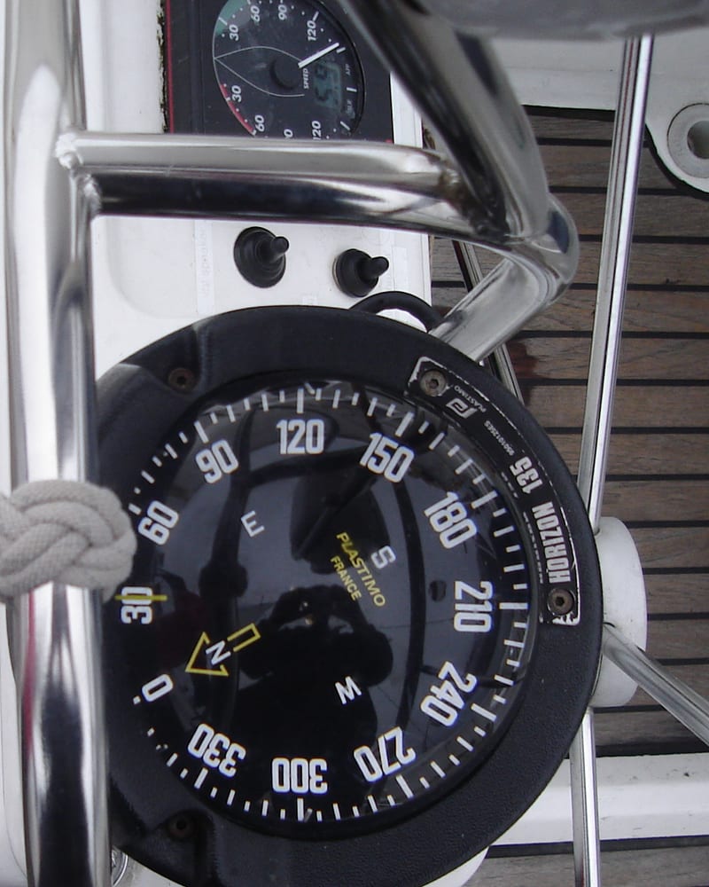 A steering compass mounted on a yacht binnacle above a grey teak deck.