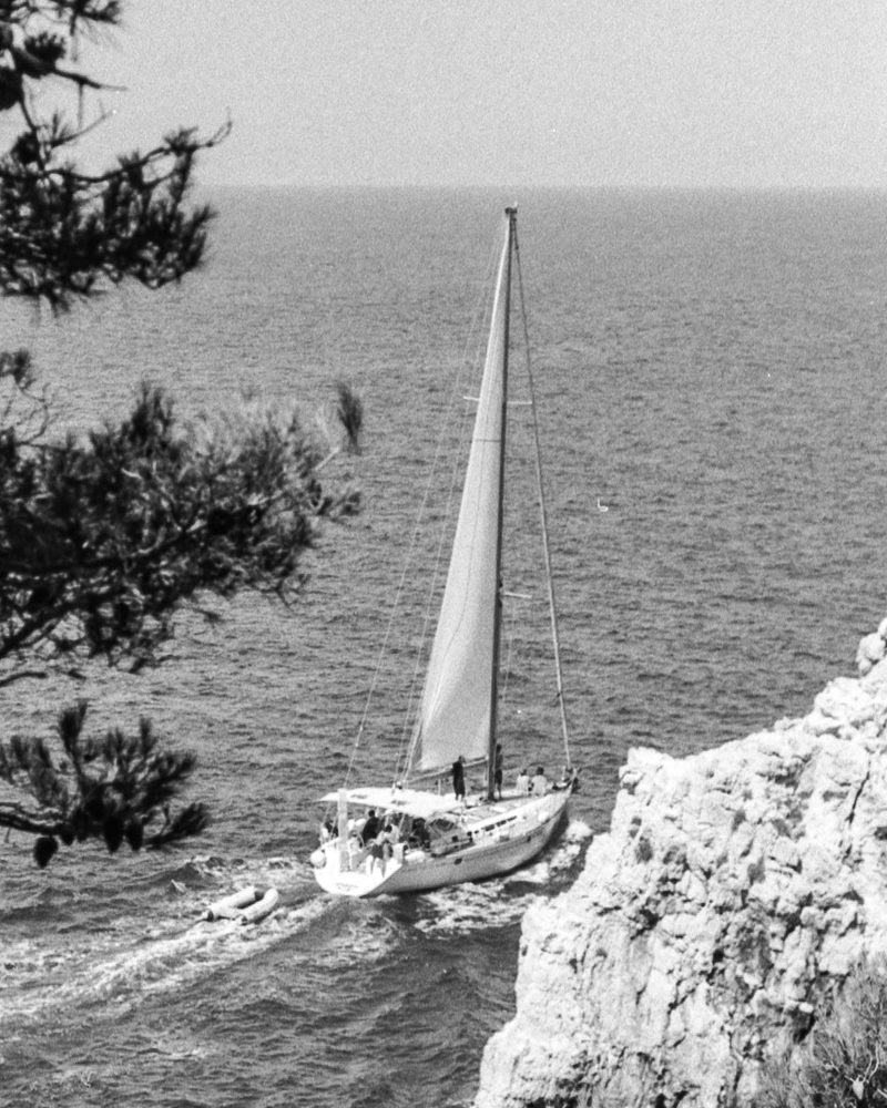 A sailing yacht under mainsail and jib, tender in tow, working close along a rocky limestone coast.