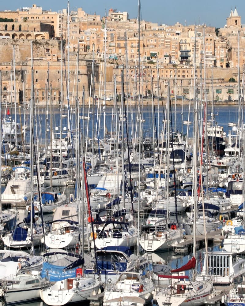 A marina crowded with yacht masts below the honey-coloured stone buildings of an old harbour city.