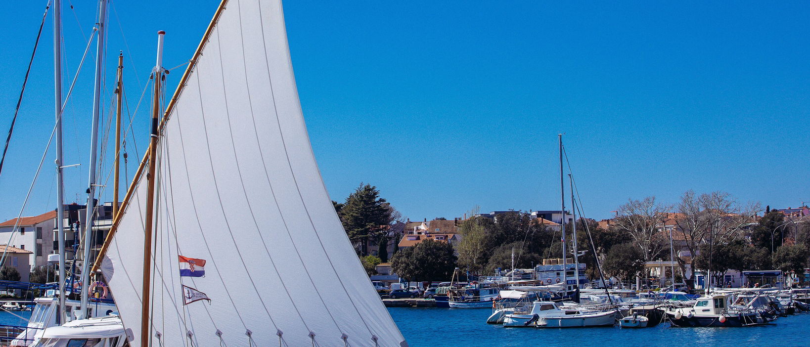 A white lateen sail in front of a harbour of moored yachts and red-roofed houses under a blue sky.